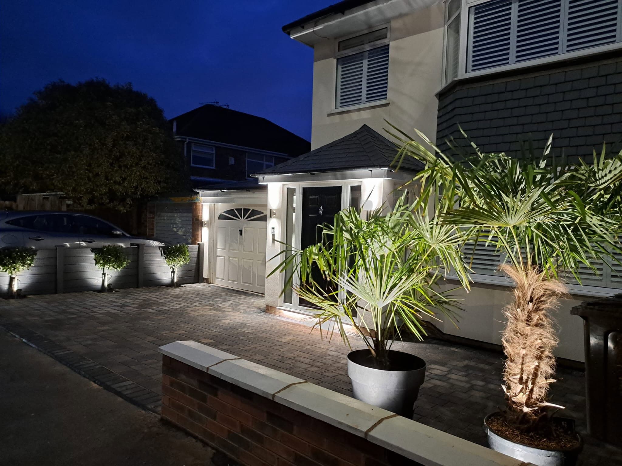 Front of a home at night with up and down wall lights, a lit porch and uplit planters