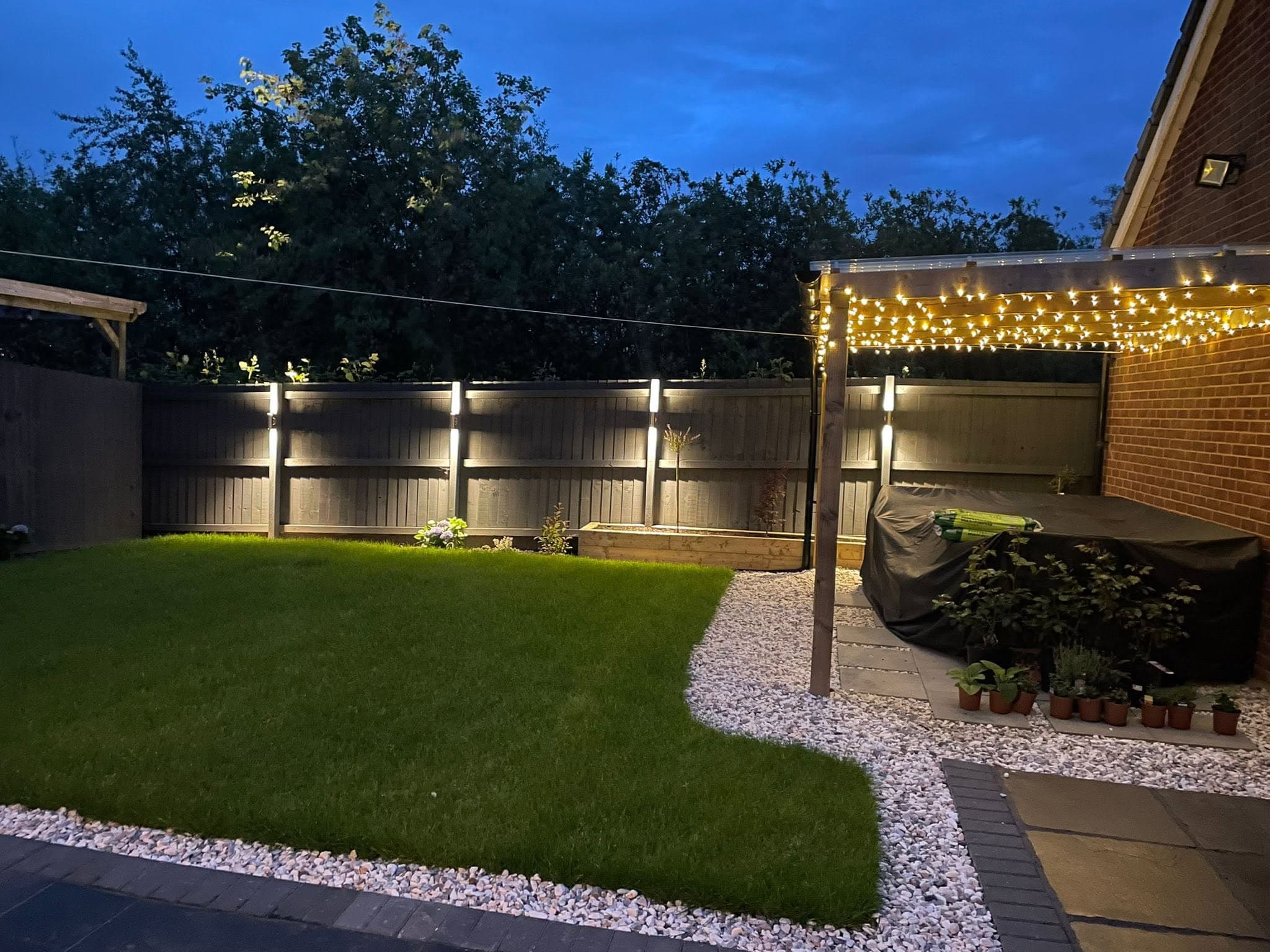 Landscaped garden at dusk lit by fence downlights and pergola festoon lighting